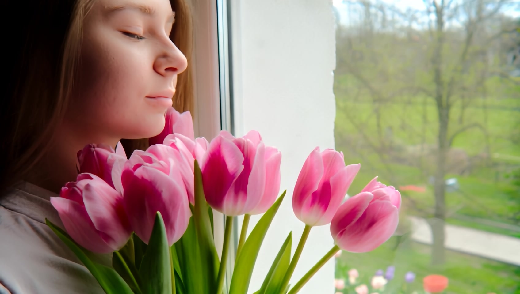 a young woman  holding bouquet of pink tulips, this young woman is hugging this bouquet of tulips, this woman is standing next to the window, behind this window is a spring blooming garden