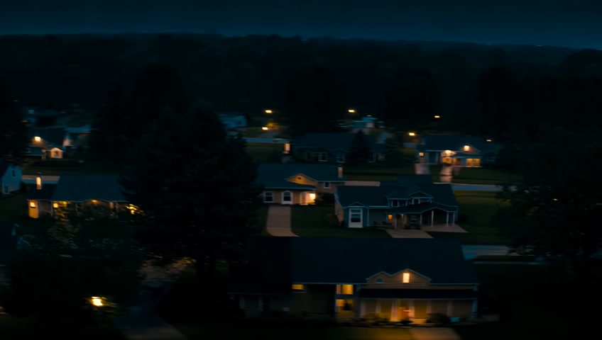 An American suburban neighborhood at night, lined with single-family homes, each with its own front yard and driveway. Some windows are illuminated with warm light, while others remain dark. Streetlights on the corners cast a dim light onto the wet sidewalk and pavement. Trees can be seen lining the street.