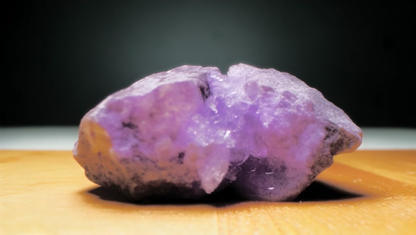 A surreal, hyperrealistic video of a lavender crystal rock sitting on a wooden surface. The rock is cracking, glowing faintly with bubbling crystals and shimmering purple goo inside. In slow motion, the rock breaks, the crystal surface crumbles and the glowing purple goo and crystal fragments from within the crystal rock spill out onto the surface, dripping and oozing in a thick, shimmering flow. The motion is smooth and cinematic. The background is minimal and dark to highlight the textures. The lava glows with orange, red, and gold sparkles. The aesthetic is dreamy, tactile, and deeply satisfying — combining ASMR-inspired motion with elemental beauty.