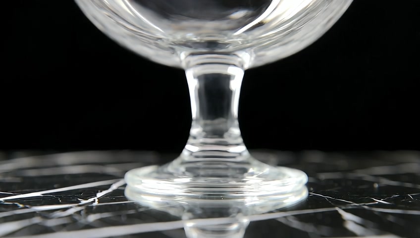 Several small, glass, hard, fruity candies are slowly poured into a large crystal punch bowl sitting on a black ceramic surface. Soft studio lighting, dark background for contrast. The candies are poured slowly, smoothly, each one landing at the bottom of the bowl.