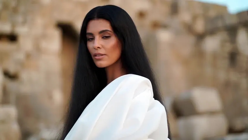 A Greek woman with olive skin and long black hair stands in front of ancient ruins, wearing a flowing white dress as she looks confidently into the camera. High resolution 4k