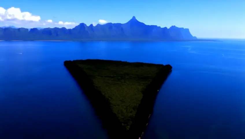 Aerial shot of a dark, triangular island covered in green vegetation surrounded by blue ocean, mountains in the distant background.