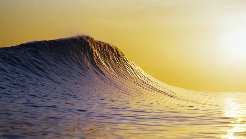 A slow-motion shot of a surfer riding a massive wave as the sun sets, casting golden hues over the ocean. The camera focuses on the surfer's silhouette against the towering wave, capturing the thrill and beauty of the moment.