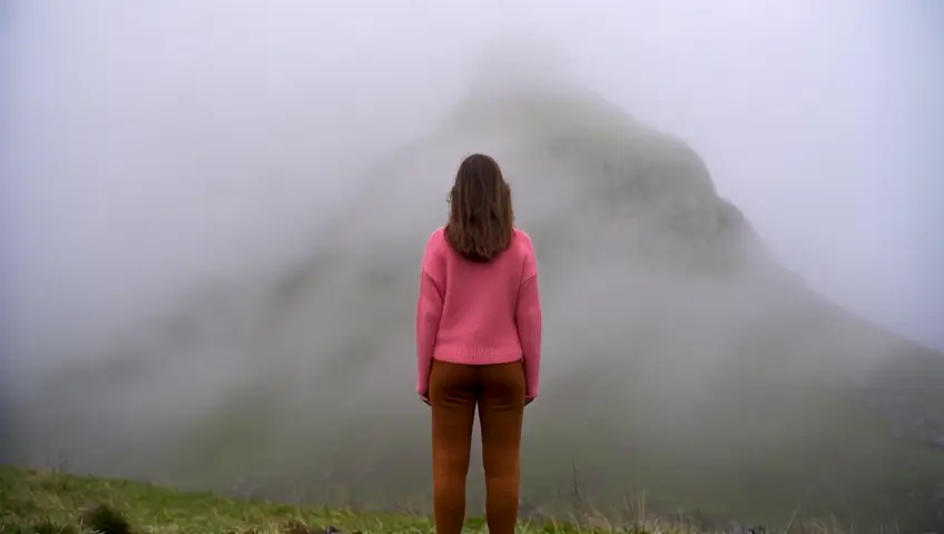 UMA MULHER CORRENDO EM DIREÇÃO A UMA MONTANHA. ELA USA BLUSA DE MALHA ROSA DE MANGA LONGA E CALÇA DE MALHA MARROM. NEBLINA