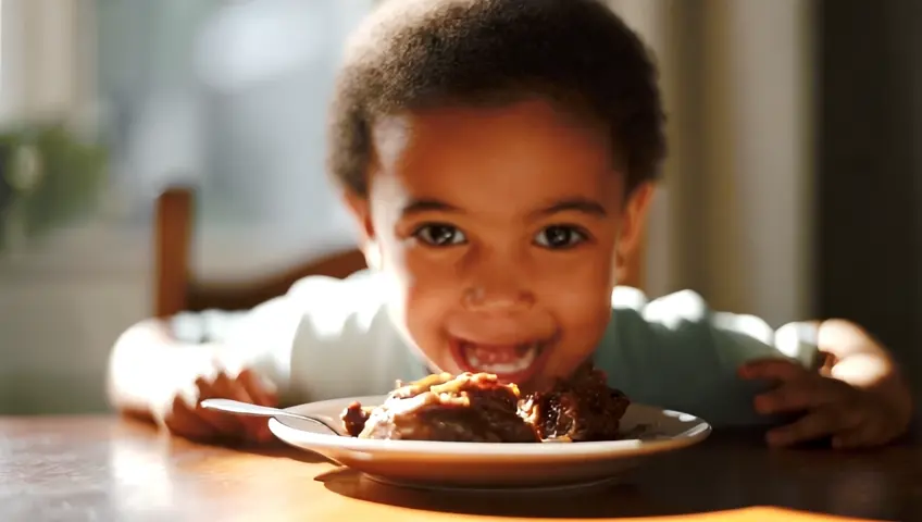 An 8-year-old child eating mutton on a plate in front of him