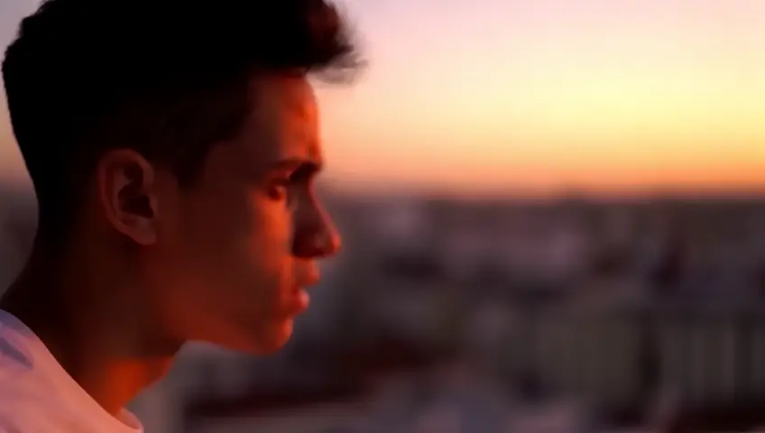 A close-up of a young man looking troubled, standing on a balcony at sunset in Madrid, with a soft breeze; vivid warm lighting captures the complex emotions of grief and shock. -ar 16:9