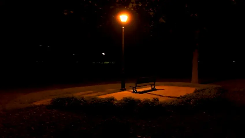 An arc shot circles a lone park bench under a streetlamp at night. The camera moves slowly, the warm light casting long shadows and creating a sense of isolation and tranquility