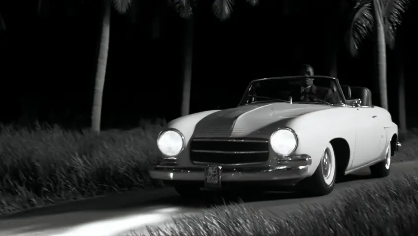 A 1950s African-Caribbean man dressed in a neatly pressed vintage suit and tie, drives a Ford Cortina along a winding Caribbean Forest road at night. his entire body and head fits in the car. The scene is illuminated by the soft silver glow of a full moon peeking through the dense canopy of towering palms and lush foliage. The car’s headlights sweep across the road, casting shifting shadows on the dense vegetation. The atmosphere is infused with a sense of nostalgia and mystery, accentuated by a grainy VHS aesthetic, black and white.