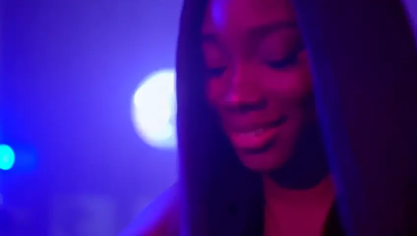 Une belle femme noire, DJ, souriante,avec des cheveux longs et lisse, mixe dans une boite de nuit, éclairée par des spots lumineux et une boule à facettes