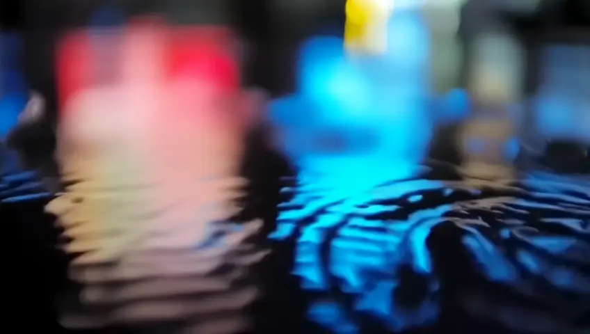 Close-up shot of raindrops falling into a puddle, with concentric ripples distorting the reflected neon signs and city lights. The camera is angled using a Dutch tilt