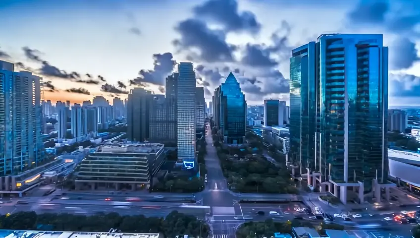 Time-lapse of a cityscape from dawn to dusk, capturing the changing light and shadows on the buildings and streets. Clouds drift overhead, and car headlights create streaks of light