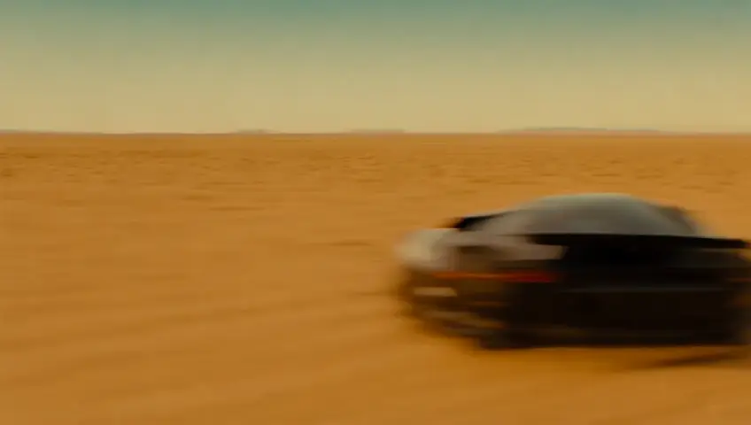 An intense, high-speed race between a black Lamborghini and a black Nissan Skyline GTR R34 across a vast desert landscape. The cars kick up clouds of dust as they tear through the barren terrain under a golden sunset. The scene alternates between close-up shots of roaring engines, spinning tires, and determined drivers, and wide-angle views capturing the endless horizon and dramatic trails of sand.