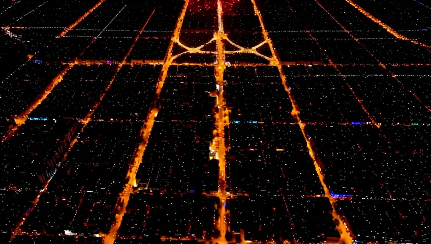 An aerial shot of a city illuminated during a festival, streets forming patterns. The camera reveals the larger design and celebration