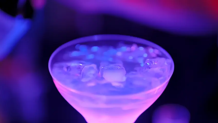 "A close-up of a drink being mixed, with liquid catching vibrant neon hues as it’s poured. Ice cubes glow faintly from the reflection of nearby LEDs. The bartender performs a flair trick, spinning the bottle mid-air as partygoers cheer in the background. Ultra-detailed focus on the drink’s surface tension and condensation drops."
