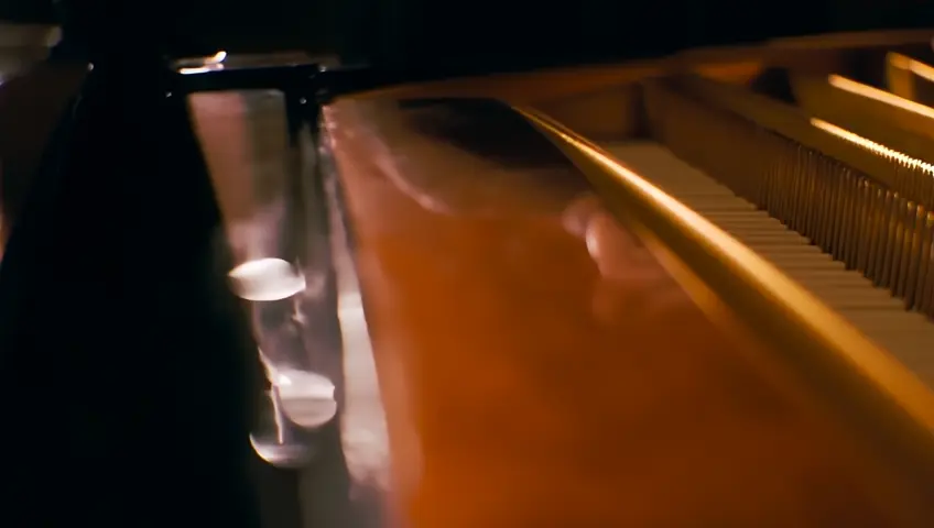 A close-up shot of a musician's hands playing a grand piano in a dimly lit concert hall. The camera focuses on the movement of the fingers and the reflection on the polished surface.