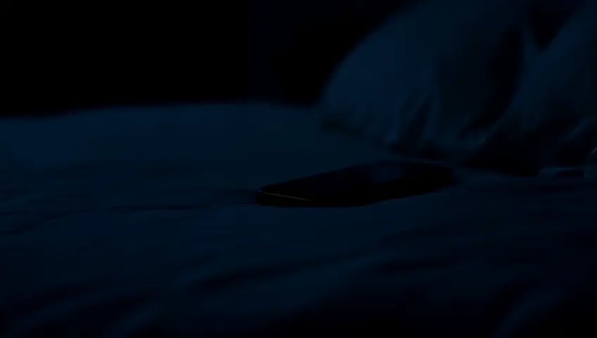 Escena realista y cinematográfica. Un celular recibe una llamada, este celular se encuentra en una cama con almohada y cobijas blancas. En el cuarto donde se encuentra la cama está con las luces apagadas, y lo que alumbra el cuarto, es la luz del celular.