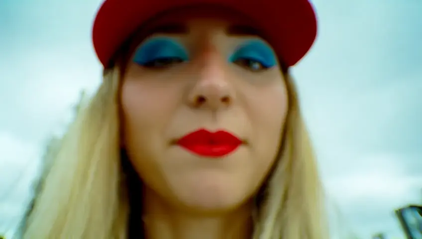 Medium close up. Low-angle shot. A teen woman with blue shadows on her eyelids, and red lipstick and a red cap. She has blonde hair. diffuse light. The person blows bubblegum pink bubble. One pops, sending tiny fragments flying. The camera zooms in on the popping bubble, capturing the intricate details