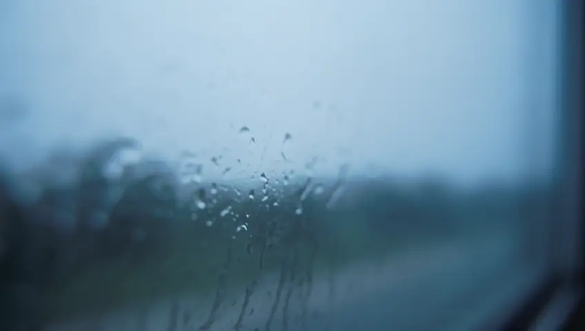 A close-up shot of rain droplets racing down a windowpane on a gloomy day. The camera focuses on the patterns and mood
