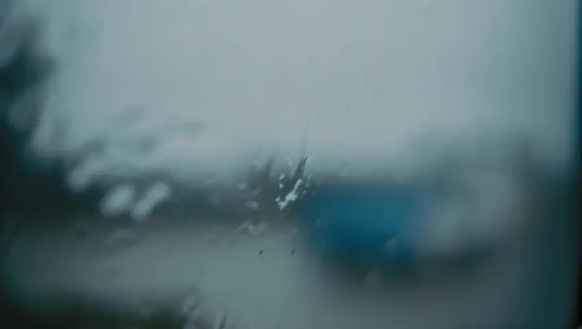 A close-up shot of rain droplets racing down a windowpane on a gloomy day. The camera focuses on the patterns and mood