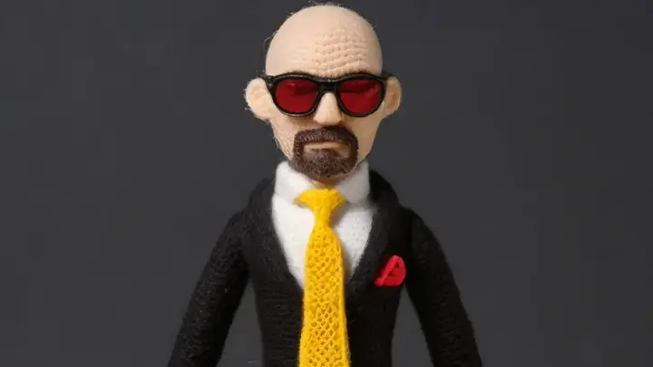 a crocheted doll of a man with a shaved head and full goatee wearing red sunglasses and a black suit with a white shirt and yellow tie