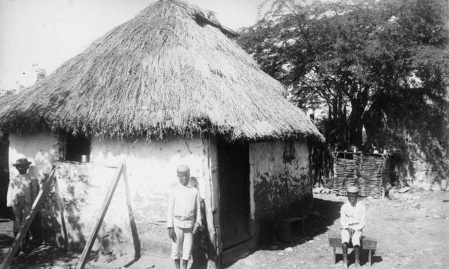 a black and white photo of an east Indian man standing in front of a hut