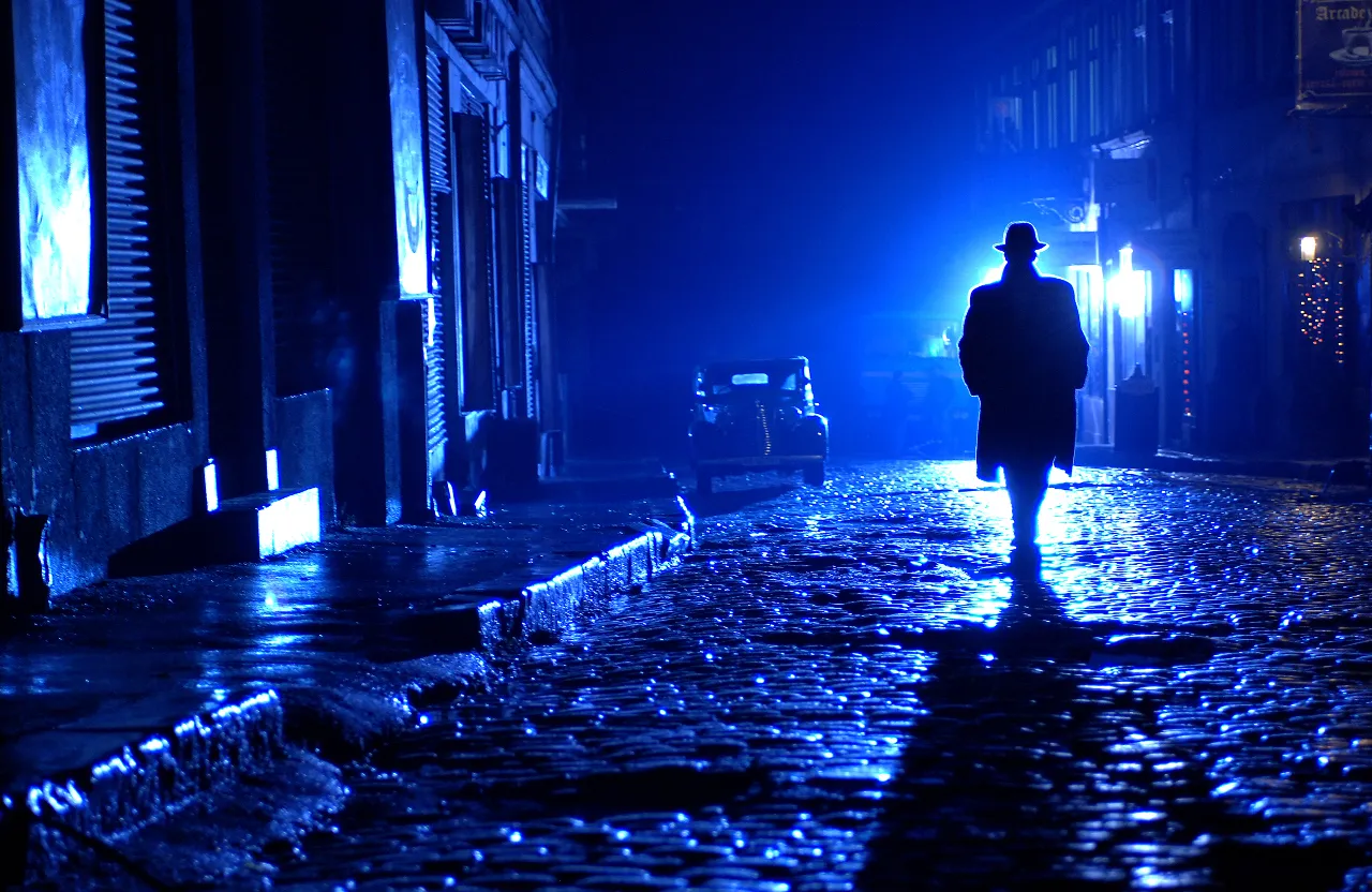 A man walking down a rainy street at night, with reflections of lights bouncing off the pavement