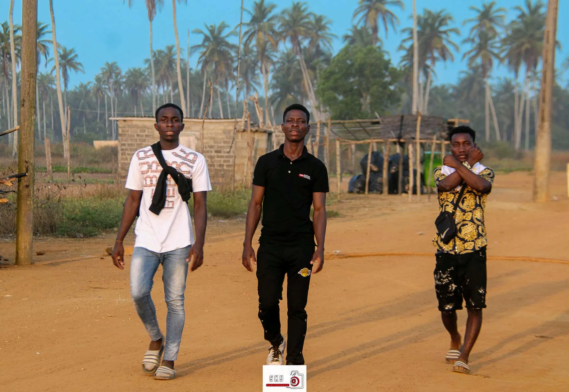 Trois jeunes hommes marchent sur un chemin de terre avec des palmiers en arrière-plan. Celui au centre porte un t-shirt noir et des chaussures de sport, tandis que les deux autres portent des vêtements plus décontractés, l'un avec un t-shirt blanc et l'autre un chemisier à motifs. Ils marchent dans un environnement rural, avec des bâtiments et des structures en arrière-plan, probablement dans une région tropicale ou côtière en raison des nombreux palmiers qui bordent la scène., advertising style