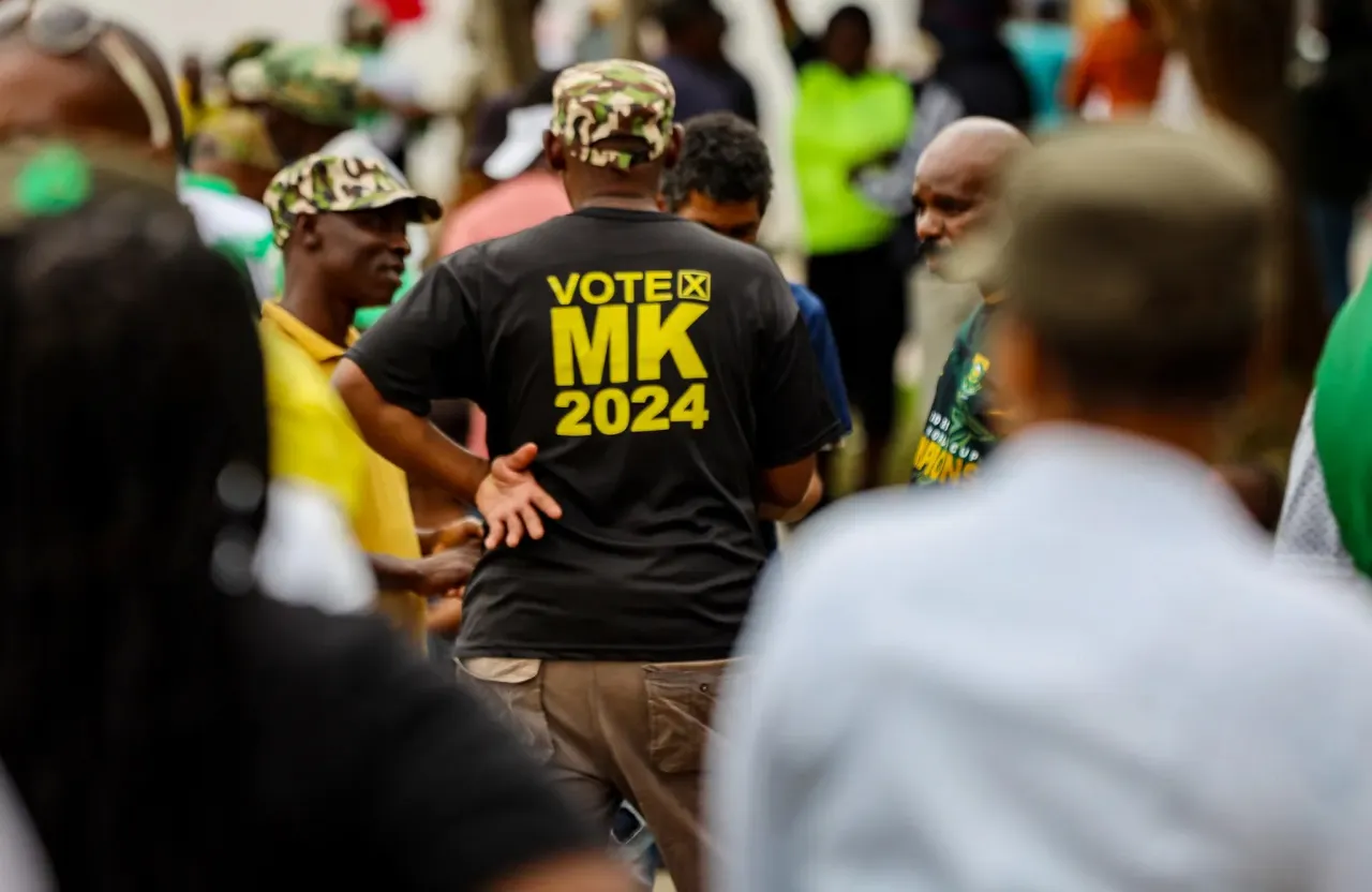 a man surrounded by a group of people. His back is facing the camera. he is wearing a black toshirt with large yellow letters MK