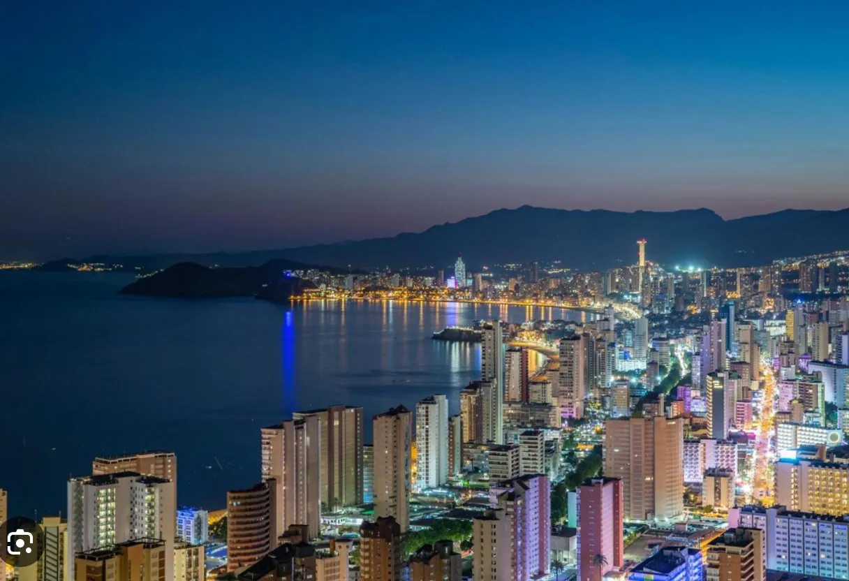 a view of a city at night from the top of a hill. No muevas los elementos de la imagen. Simplemente anima la cámara como un dron. Añade carteles de Neón gigantes estáticos entre los edificios. Escribe en uno de ellos ''Benidorm''