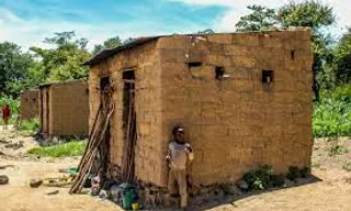 dilapidated hut at the edge of the village in Africa, its walls patched together with scraps of wood, plastic and metal sheeting, clean outside, people living in it, 2 children playing outside, woman washing utensils, small door, a charcoal pit nearby