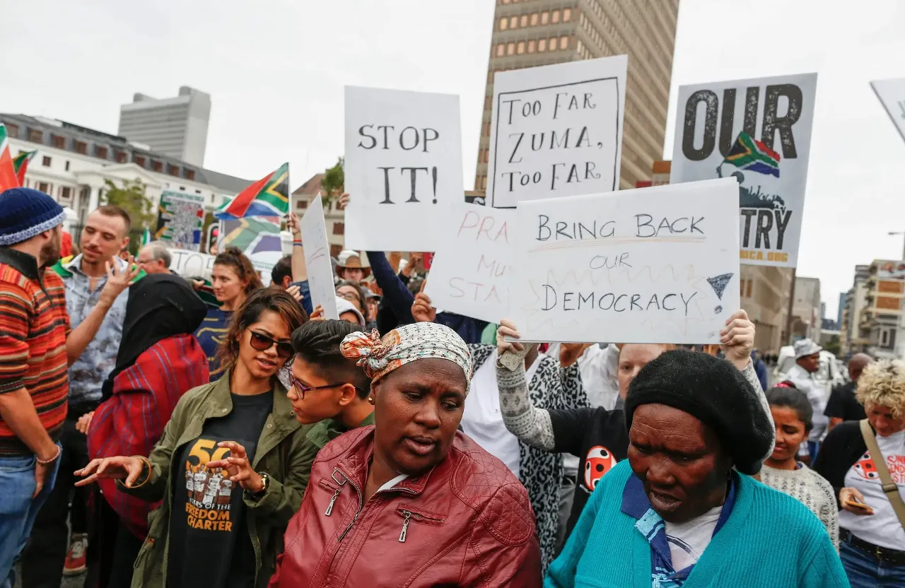 a group of people holding signs in a protest. They hold a placard that says 'too far Zuma! Too far!