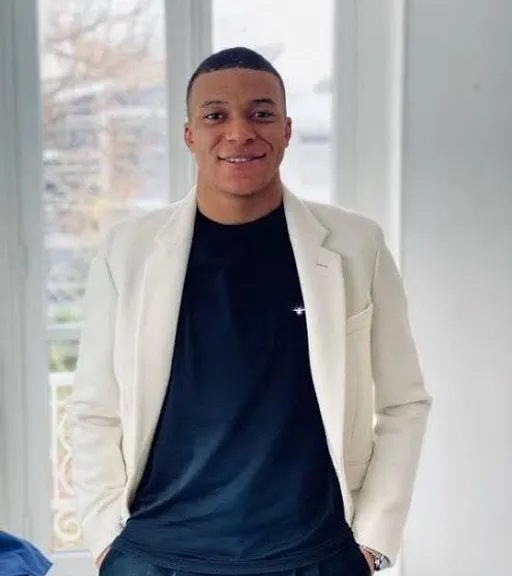 Mbappe in a white blazer standing in front of a window