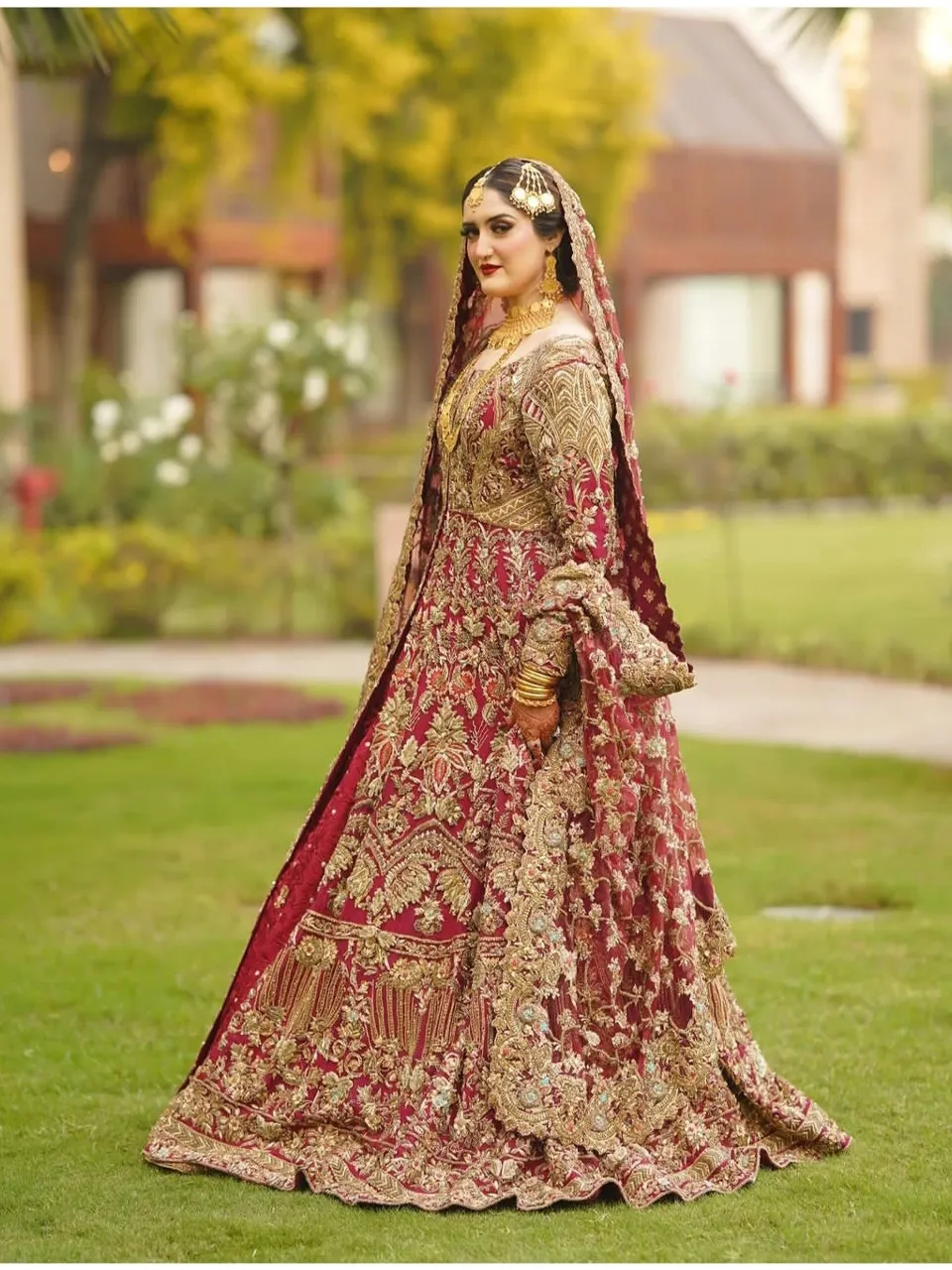 A bride in a red and gold gown surrounded by blooming cherry blossoms under a canopy of trees during sunset