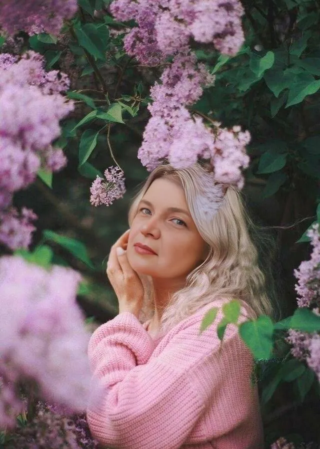 a woman in a pink sweater standing in front of purple flowers the movement of flowers from the wind