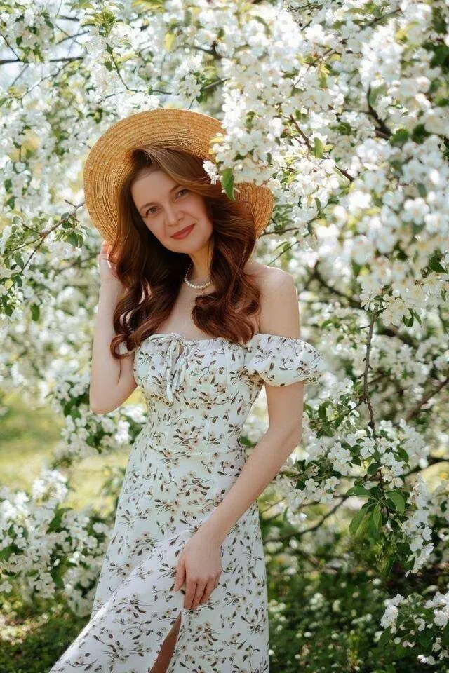 a woman in a dress and straw hat standing in front of a flowering tree the movement of flowers from the wind