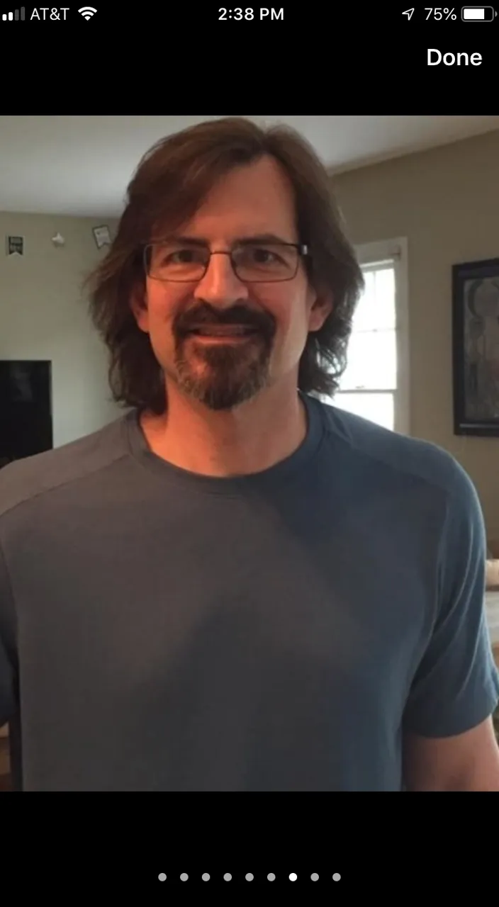 Long haired man with mandrake style facial hair and fit body stares into the camera while buildings collapse all around him