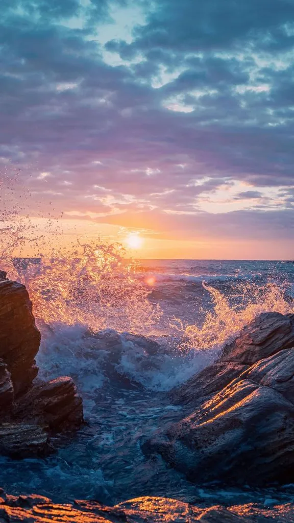 the sun is setting over the ocean with waves crashing on the rocks