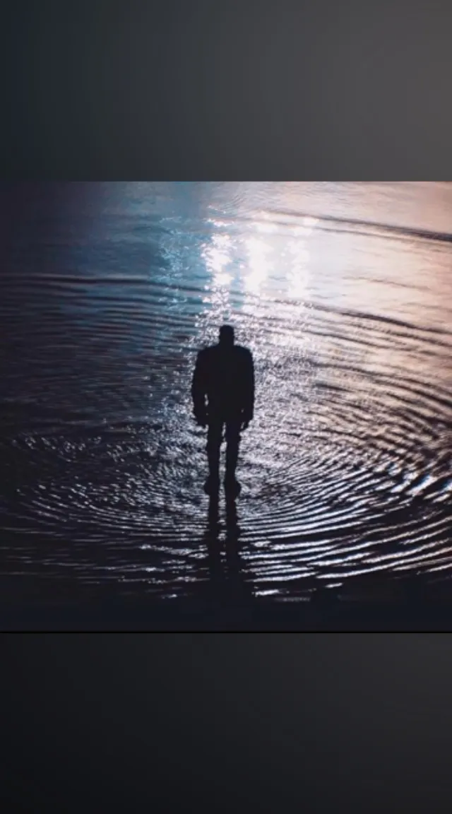 a shadow silhouette of a man thinking while standing on a body of water in the center of gentle ripples that shimmer with light and color